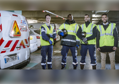 Équipe de techniciens en gilets haute visibilité dans un parking souterrain, debout à côté d’un fourgon de service marqué "SDEL Transport Services", équipé de bandes réfléchissantes et de signalisation de sécurité.